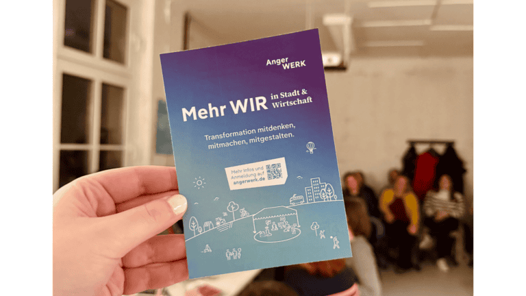 Hand hält Flyer mit "Mehr WIR" in einem Raum mit Menschen im Hintergrund.