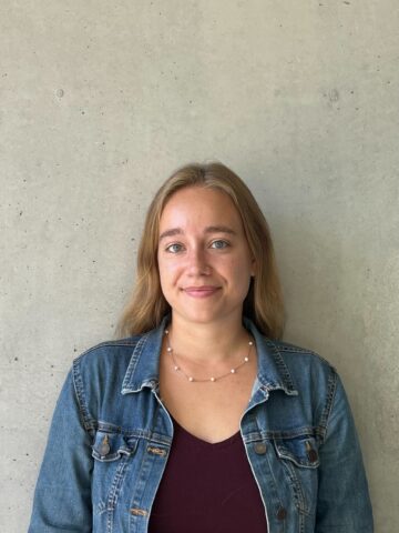 Junge Frau mit blonden Haaren und Jeansjacke vor Betonwand.