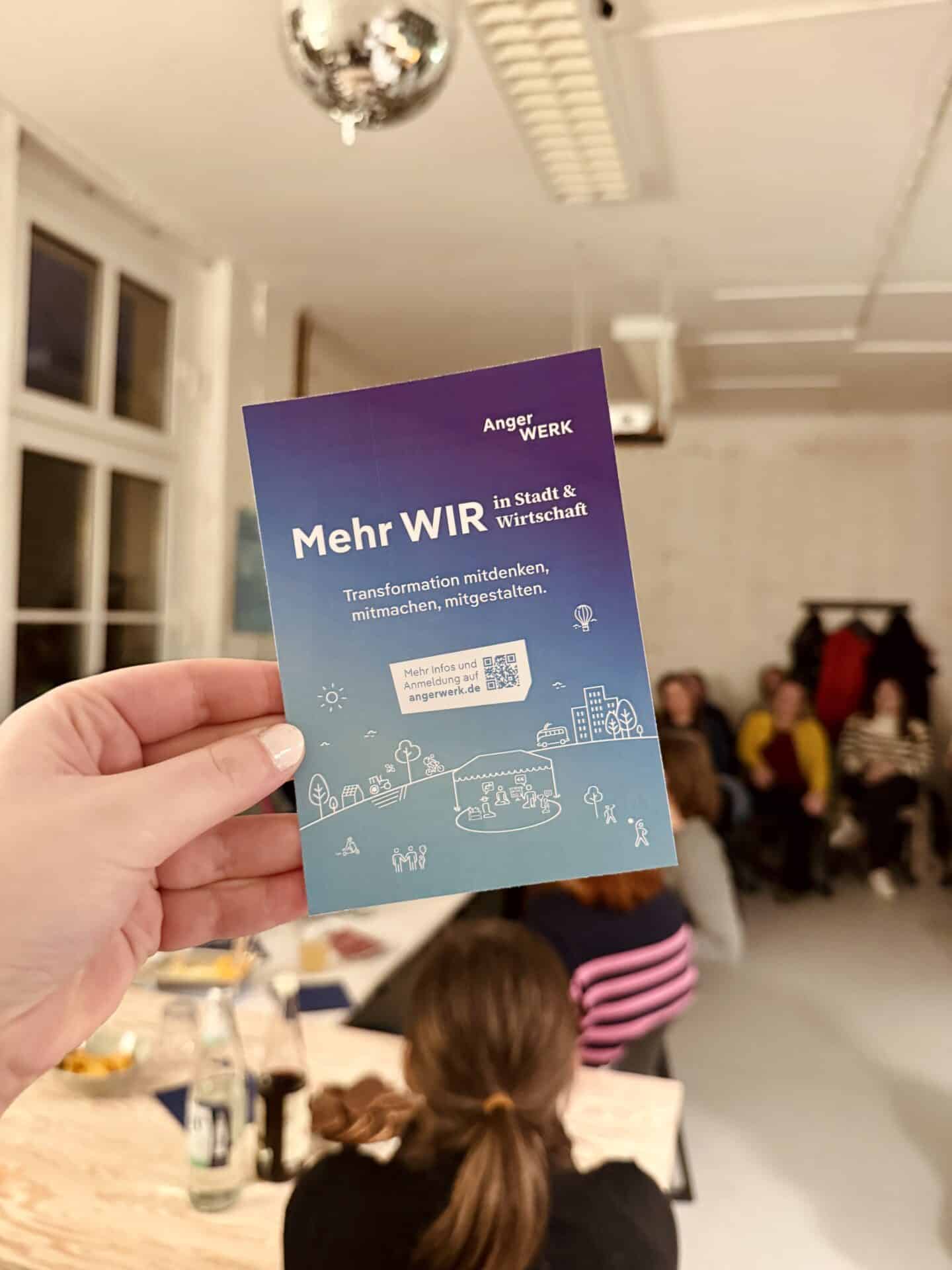Hand hält Flyer mit "Mehr WIR" in einem Raum mit Menschen bei einem Event.