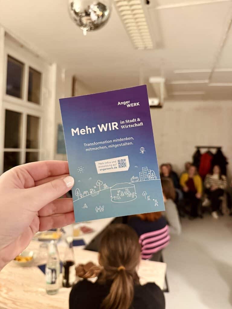 Hand hält Flyer mit "Mehr WIR" in einem Raum mit Menschen bei einem Event.