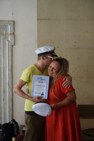 Zwei Menschen umarmen sich, einer hält ein Zertifikat, im Hintergrund eine weiße Mütze.