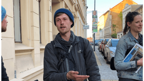 Junger Mann in dunkler Jacke und Mütze auf einer Stadtstraße, spricht mit jemandem.