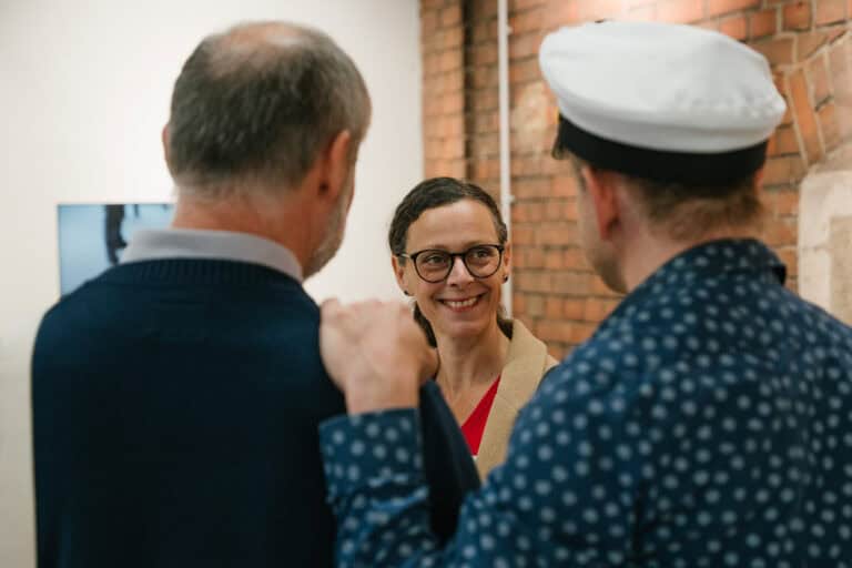 Drei Menschen im Gespräch, eine Frau lächelt, im Hintergrund eine Ziegelwand.