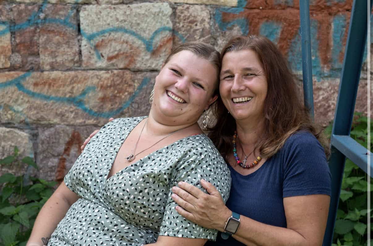 Freundinnen lachen im Garten vor einer Steinmauer mit Graffiti, genießen den Moment und zeigen ihre.